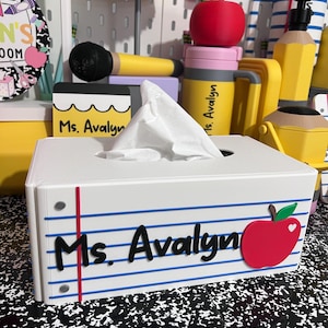 May include: A rectangular white tissue box cover designed to resemble a piece of lined notebook paper. It features blue horizontal lines, a vertical red margin line, the black text Ms. Avalyn, and a red apple graphic with a green leaf on the front.