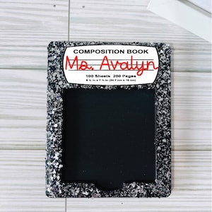 May include: Black composition book with a silver glitter border. The front cover has a white label with the text "COMPOSITION BOOK" and "Ma. Avalyn" written in red. The label also includes the text "100 Sheets 200 Pages" and "9 1/4 in x 7 1/2 in (24.7 cm x 19 cm)".