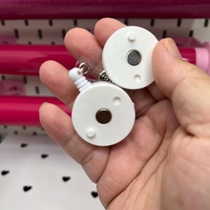 May include: Two white, round badge reel attachments with a magnetic backing. The badge reels have a retractable cord and a clear plastic strap. The background includes pink rolls of paper and a white surface with black heart shapes.