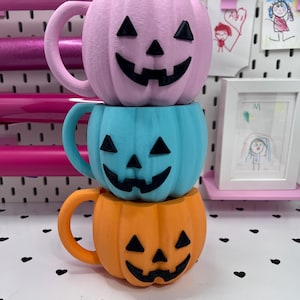 May include: A stack of three pumpkin-shaped mugs, each featuring a black jack-o'-lantern face. The top mug is light pink, the middle mug is light blue, and the bottom mug is orange. Each mug has a curved handle on the left side.