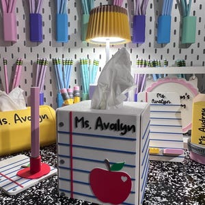 May include: A white square tissue box cover designed to look like a piece of notebook paper with blue horizontal lines, a red vertical margin, and a red apple cutout. The text Ms. Avalyn is printed on the front. A small lamp sits on top.