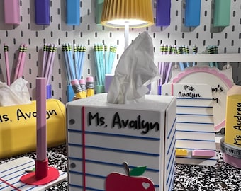May include: A white square tissue box cover designed to look like a piece of notebook paper with blue horizontal lines, a red vertical margin, and a red apple cutout. The text Ms. Avalyn is printed on the front. A small lamp sits on top.