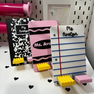 May include: Three custom phone stands shaped like school supplies. One is a black and white composition notebook, one is a pink crayon, and one is a piece of lined notebook paper. Each stand features the name Ms. Avalyn and a yellow support ledge.