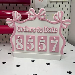 Può includere: Un cartello decorativo bianco e rosa con la scritta "Orders to Date" e il numero "8557". Il cartello presenta un design a fiocco e si trova su una superficie bianca con decorazioni a forma di cuore nero.