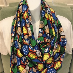 May include: A colorful infinity scarf featuring a repeating pattern of comic book heroes. The design includes characters in red, blue, green, and yellow on a black background. The scarf is displayed on a white mannequin.