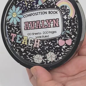 May include: A round, black container with a composition book design. The design includes a flower, pencil, rainbow, and the text "COMPOSITION BOOK AVALYN TEACH 100 Sheets 200 Pages Wide Ruled". The container is held in a hand.