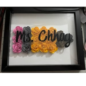 May include: A framed piece of art with the name "Ms. Chhay" written in black lettering. The background is made of paper flowers in pink, gray, and yellow.
