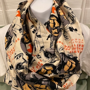 May include: A cream-colored infinity scarf with a black and orange print of a comic book style illustration of soldiers and military vehicles. The text on the scarf reads "MARIF SHORE DEF BATTALION", "VEOZ", and "NS".