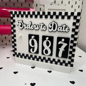 May include: A white and black checkered "Orders to Date" counter with the number "987" displayed. The counter is made of acrylic and sits on a white surface with black heart-shaped confetti. The background includes rolls of pink vinyl.