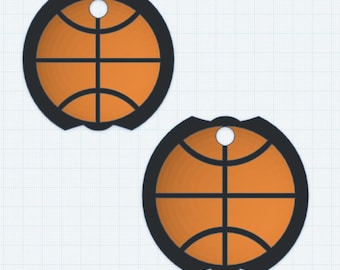 May include: Two orange and black basketball-themed pendants. Each pendant features a basketball design with black lines and a black border. A small hole is at the top for hanging. The pendants are set against a grid background.