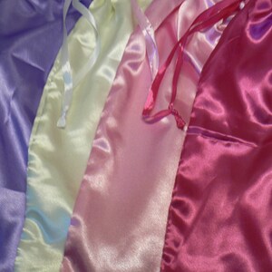 May include: A selection of satin drawstring bags in pastel shades: lavender, cream, light pink, and hot pink. Each bag features a matching satin ribbon drawstring. The bags are displayed together, highlighting their smooth, glossy finish.
