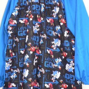 May include: A blue and black long-sleeved smock with a Sonic the Hedgehog print. The print features Sonic, Knuckles, and Tails running with the words "Let's Go" repeated throughout.