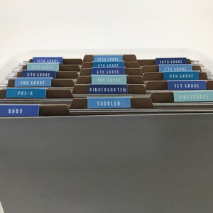 May include: A clear plastic file box with brown file folders labeled with blue and light blue labels for different grade levels, including baby, toddler, preschool, 1st grade, kindergarten, 2nd grade, 3rd grade, 4th grade, 5th grade, 6th grade, 7th grade, 8th grade, 9th grade, 10th grade, 11th grade, and 12th grade.