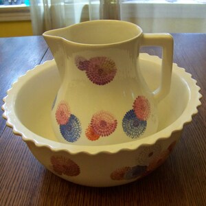 Könnte beinhalten: Cremefarbenes Krug- und Waschbecken-Set. Der Krug hat einen Henkel und ist mit floralen Mustern in Rosa, Blau und Orange verziert. Das Waschbecken hat einen gewellten Rand.