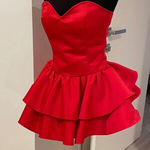 Op de afbeelding: Een felrode strapless jurk op een zwarte mannequin. De jurk heeft een sweetheart halslijn en een gelaagde, gerimpelde rok. De jurk is gemaakt van een glanzend, satijnachtig materiaal en staat op een zilveren metalen standaard.
