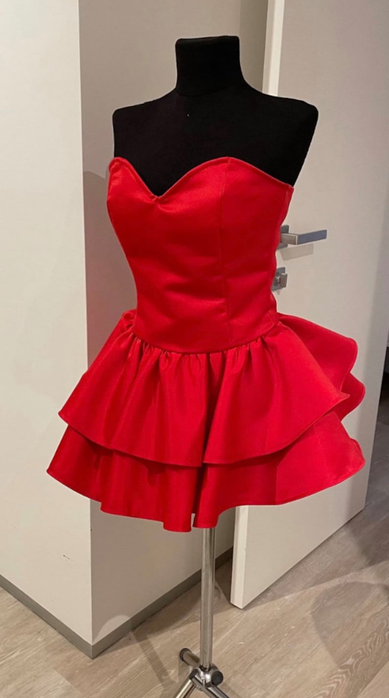 Op de afbeelding: Een felrode strapless jurk op een zwarte mannequin. De jurk heeft een sweetheart halslijn en een gelaagde, gerimpelde rok. De jurk is gemaakt van een glanzend, satijnachtig materiaal en staat op een zilveren metalen standaard.