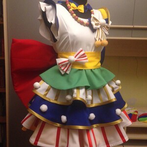 Op de afbeelding: Een kleurrijk cosplaykostuum op een mannequin. De outfit heeft een witte top, een gelaagde rok met groene, blauwe en witte secties en rood-witte streepaccenten. Het bevat een grote rode strik, gele accenten en decoratieve accessoires.