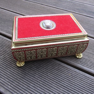 May include: A red velvet-lined box with gold trim and ornate detailing. The box has a raised, oval-shaped design on the lid with a white cameo.