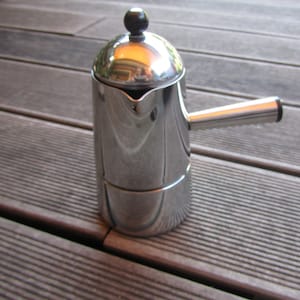 Può includere: Una macchina per caffè espresso in acciaio argentato con una manopola nera sul coperchio. La macchina è appoggiata su una superficie di legno.