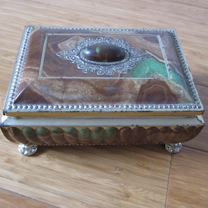 May include: A vintage metal tin box with a decorative lid. The lid features a brown and green marbled design with a silver-colored ornate embellishment and a green gem-like center. The box has a silver-colored trim and sits on four small feet.