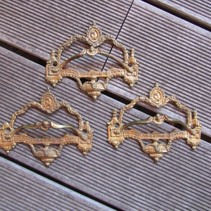 Op de afbeelding: Drie sierlijke, goudkleurige metalen ladegrepen met ingewikkelde ontwerpen. Elke greep heeft een gebogen handvat en een decoratieve bovenkant.