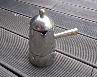 May include: A silver stainless steel stovetop espresso maker with a gold knob on the lid. The maker is sitting on a wooden surface.