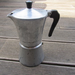 May include: A silver aluminum stovetop coffee maker with a black handle and lid. The coffee maker is sitting on a wooden surface.