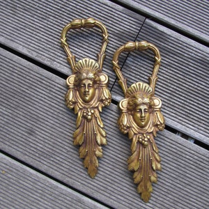 May include: Two gold-toned metal decorative wall hooks with a stylized female face design. Each hook features a detailed floral and leaf motif.