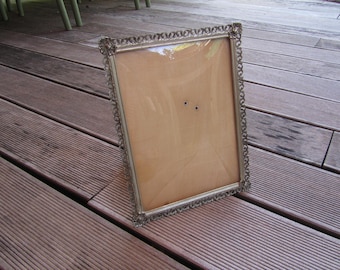 May include: An ornate, rectangular picture frame with a decorative border. The frame is made of a gold-colored metal and has a light brown backing. The frame is empty and ready for a photo or artwork.