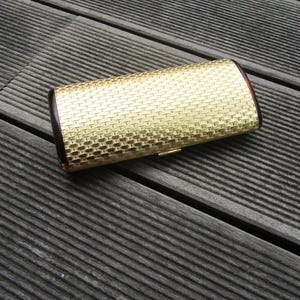 Op de afbeelding: Een gouden clutch met een geweven textuur. De clutch heeft een bruine rand.