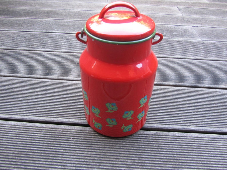 May include: Red enamel milk can with a green lid and floral pattern. The can has a handle on the top and is sitting on a wooden surface.