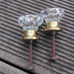 Puede incluir: Dos pomos de puerta de vidrio transparente con bases de latón y tornillos de metal oxidado. Los pomos tienen un diseño facetado.