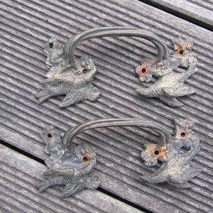 Peut inclure: Quatre poignées de tiroir antiques en métal. Chaque poignée présente un motif d'oiseau avec une poignée incurvée. Les oiseaux sont détaillés avec des ailes et des accents floraux, et ont un aspect vieilli et patiné. Les poignées sont montées sur une surface en bois.