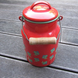 May include: Red enamel milk can with a wooden handle and a floral design. The can has a green rim and a shiny finish.