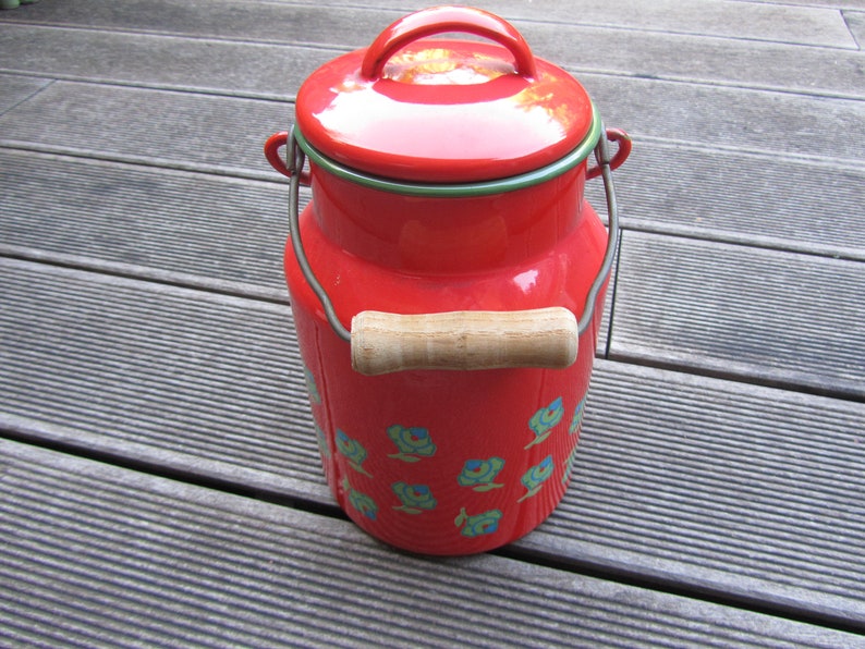 May include: Red enamel milk can with a wooden handle and a floral design. The can has a green rim and a shiny finish.