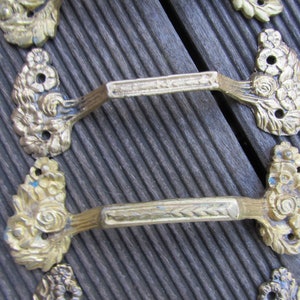 May include: Antique brass drawer pulls with floral and leaf detailing. The handles feature a rectangular centre with a textured pattern. The pulls are mounted on a dark wooden surface.