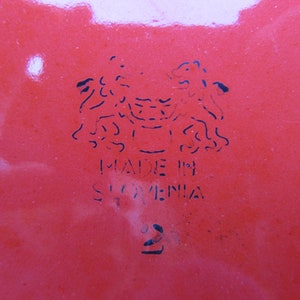 May include: A red enamelware dish with a black stamp that reads "Made in Slovenia" and a stylized image of two lions.