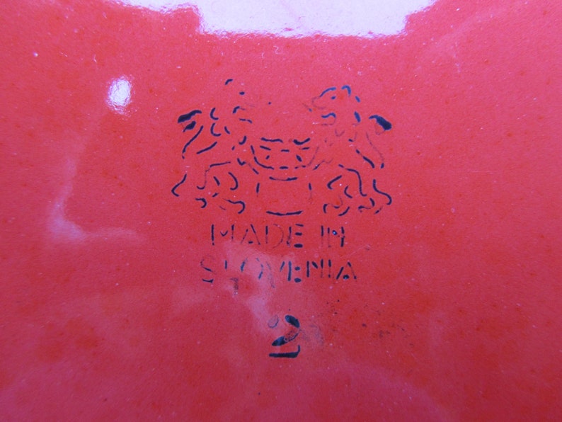 May include: A red enamelware dish with a black stamp that reads "Made in Slovenia" and a stylized image of two lions.