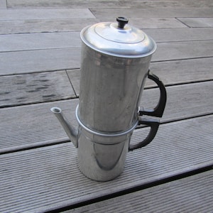 Puede incluir: Una cafetera percoladora plateada vintage con tapa y asas negras. La cafetera tiene un pico y es de metal. La cafetera está sobre una superficie de madera.