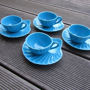 May include: Four blue ceramic teacups and saucers with a shell-like design. The cups and saucers are arranged on a wooden surface.