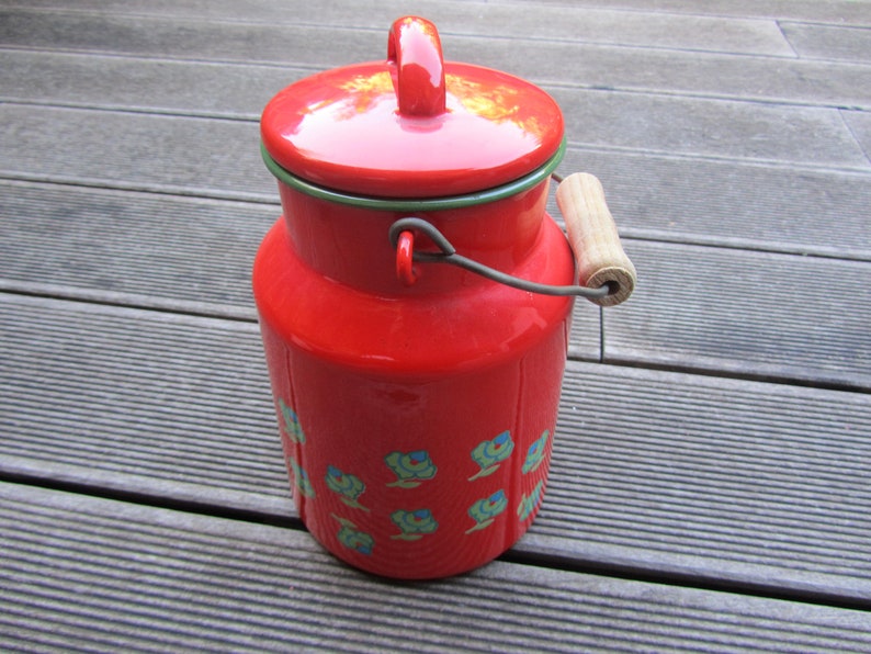 May include: Red enamelware milk can with a wooden handle and a lid. The can has a floral design in blue and green.