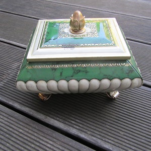 May include: A green and gold decorative tin box with a scalloped edge and three gold feet. The lid has a gold finial in the shape of a flower bud.