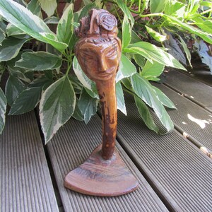 May include: A wooden sculpture of a person's head with a detailed face and a crown of leaves. The sculpture is on a wooden base and is standing on a wooden deck.