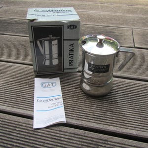 May include: A silver stainless steel coffee maker with a black label that reads "PRATIKA AUTOMATICA ESPRESSO", and a white box with the text "la caffettiera espresso" and "GAT".