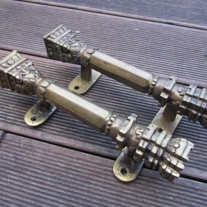 May include: Two ornate brass door handles with intricate designs. The handles are shaped like columns with decorative details and are mounted on a wooden surface.