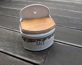 May include: White ceramic salt container with a wooden lid and a fruit design. The container has a hanging hook and says "SALE" on the front.