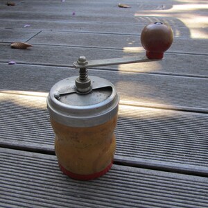 Può includere: Un macinacaffè vintage in legno con un coperchio argentato e una manopola rossa. Il macinacaffè ha una manovella e un meccanismo a molla.