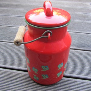 May include: Red enamel milk can with a wooden handle and a green rim. The can has a floral design with blue and green flowers.