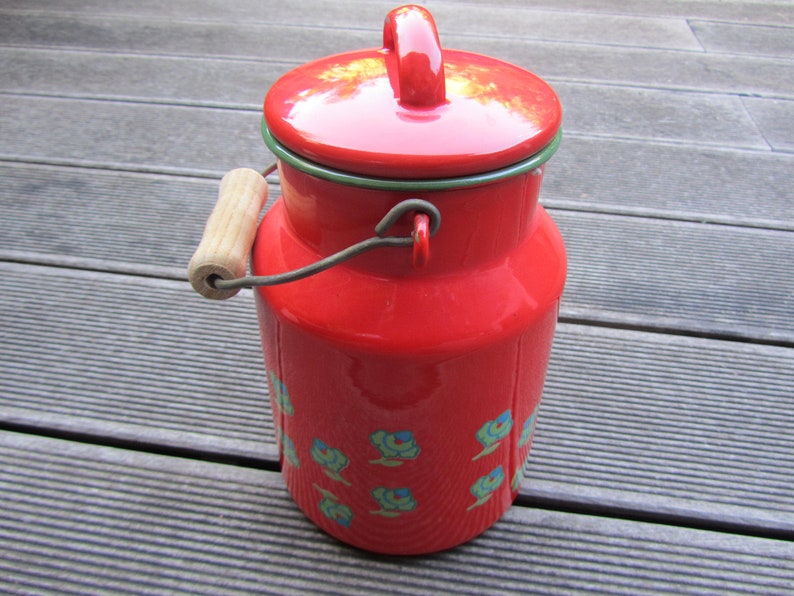 May include: Red enamel milk can with a wooden handle and a green rim. The can has a floral design with blue and green flowers.