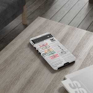 May include: A white phone case with a newspaper print design. The case has a black camera lens and a black bat logo with the text "RIN" printed on it. The case also has the text "Healing Hands" printed on it.